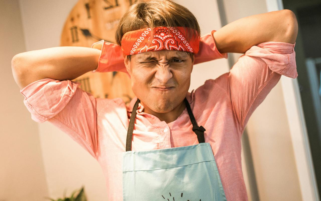 Osoba u roze košulji i plavoj kecelji pravi nervoznu grimasu sa podignutim rukama